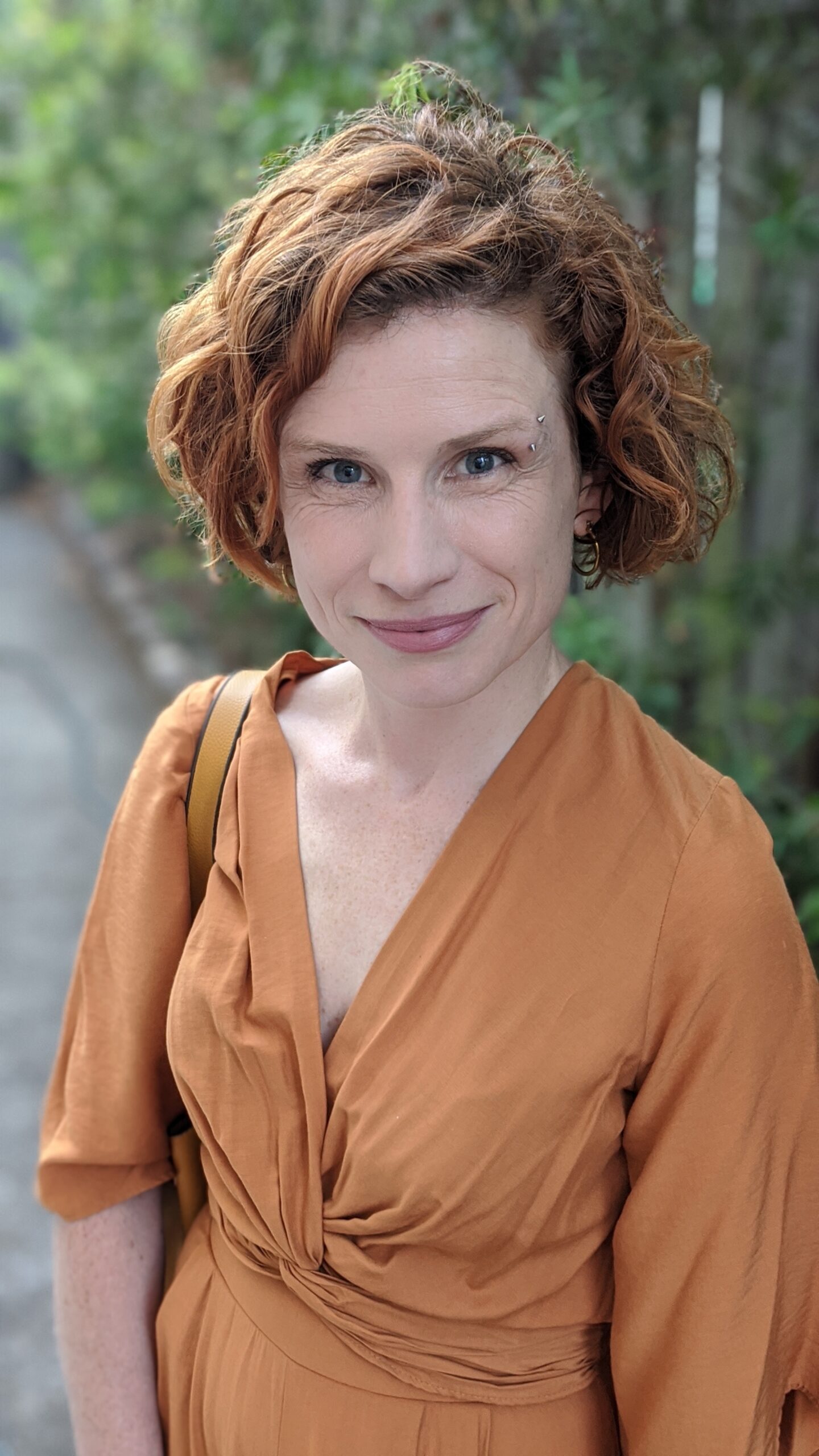 A portrait photograph of Fiona Wright. Fiona has auburn-coloured curly hair and is wearing a terracotta coloured dress.