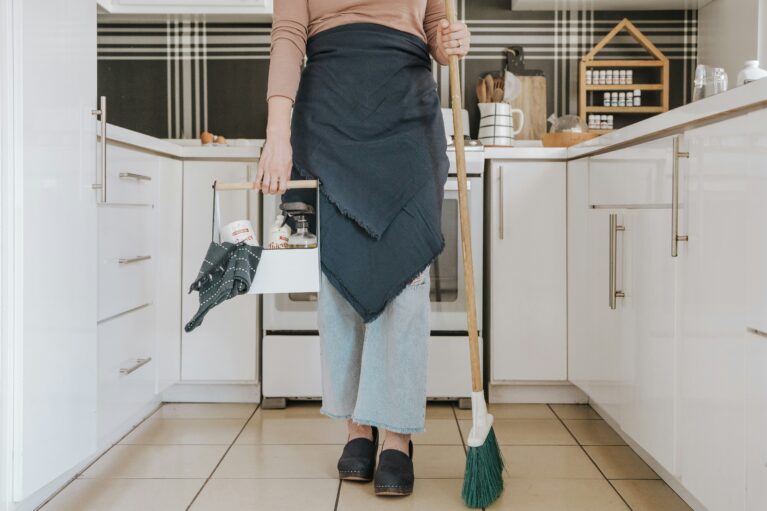 A portrait photograph of a person's torso. The person is wearing an apron. They hold a broom in one hand and a basket of cleaning products in the other.