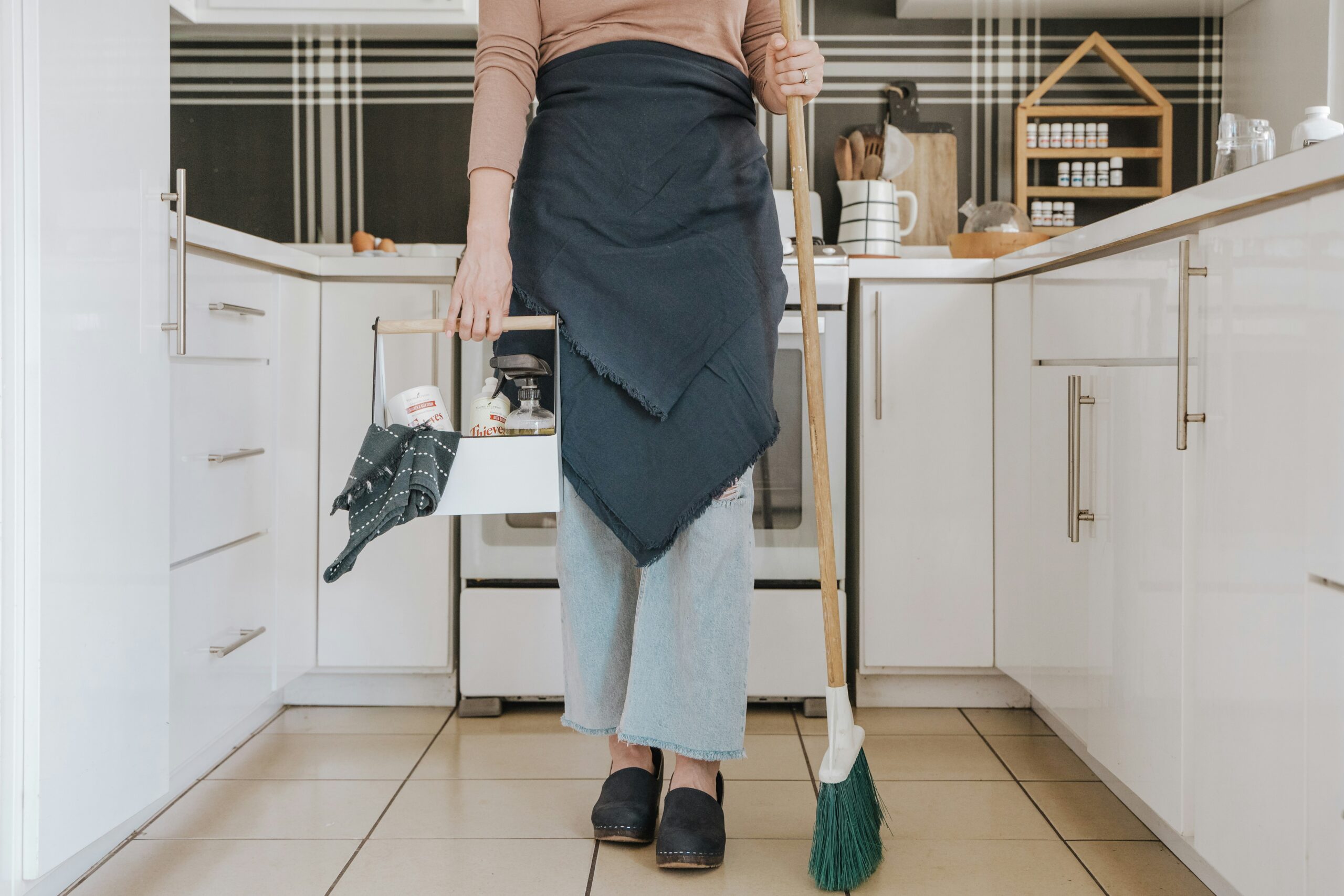 A portrait photograph of a person's torso. The person is wearing an apron. They hold a broom in one hand and a basket of cleaning products in the other.