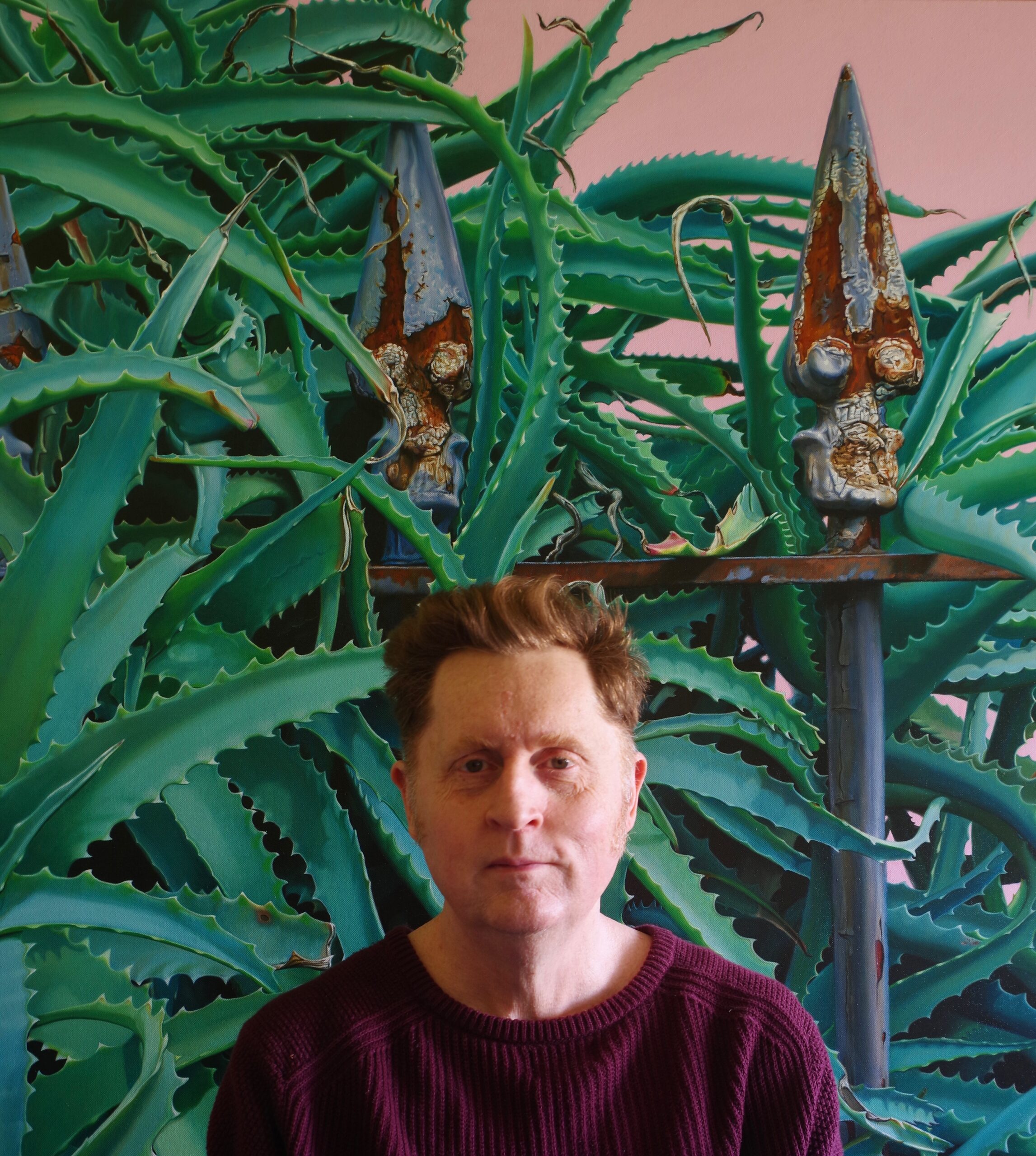 A photograph of Jason Moad. The artist is wearing a maroon sweater and standing in front of one of his paintings. The painting depicts an unruly, overgrown aloe plant growing over a rusty, wrought-iron fence.