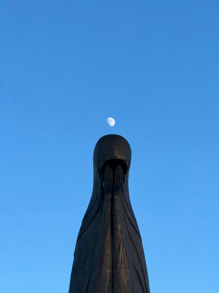 A statue in front of a bright blue sky. It has its face buried in its hands as if it is weeping.