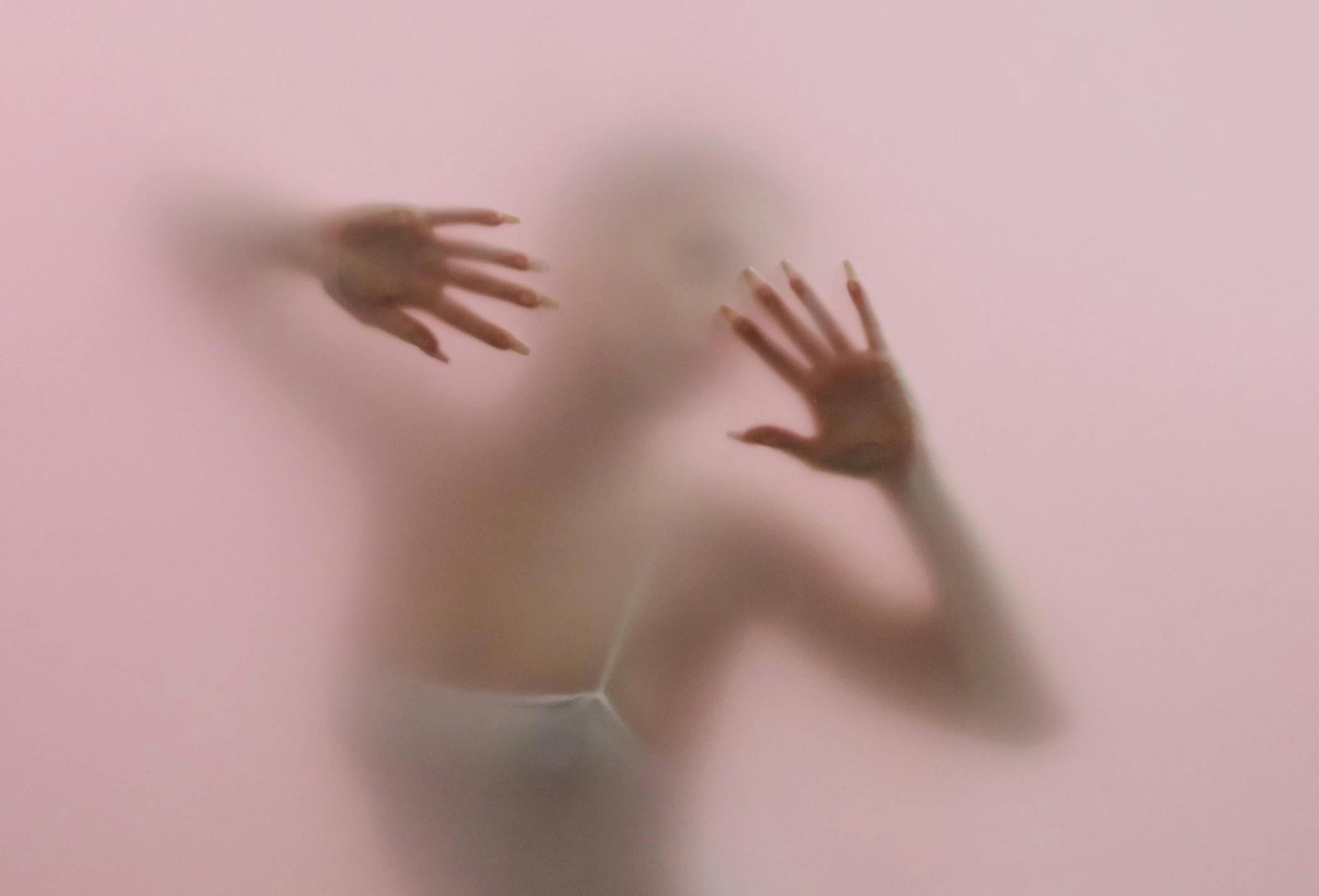 A woman's upper body and hands are visible through a fogged mirror on a pink background
