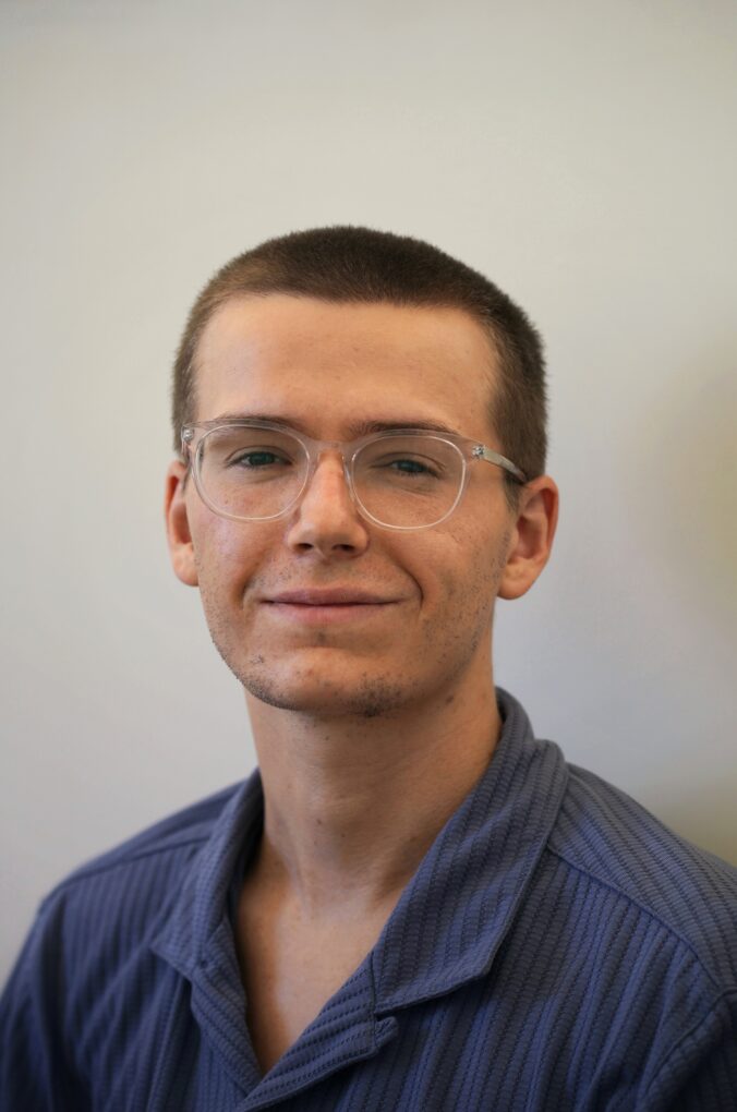 A photograph of Ned Lupson. He is smiling. His dark blonde hair is shaved. He is wearing glasses and a blue collared shirt.