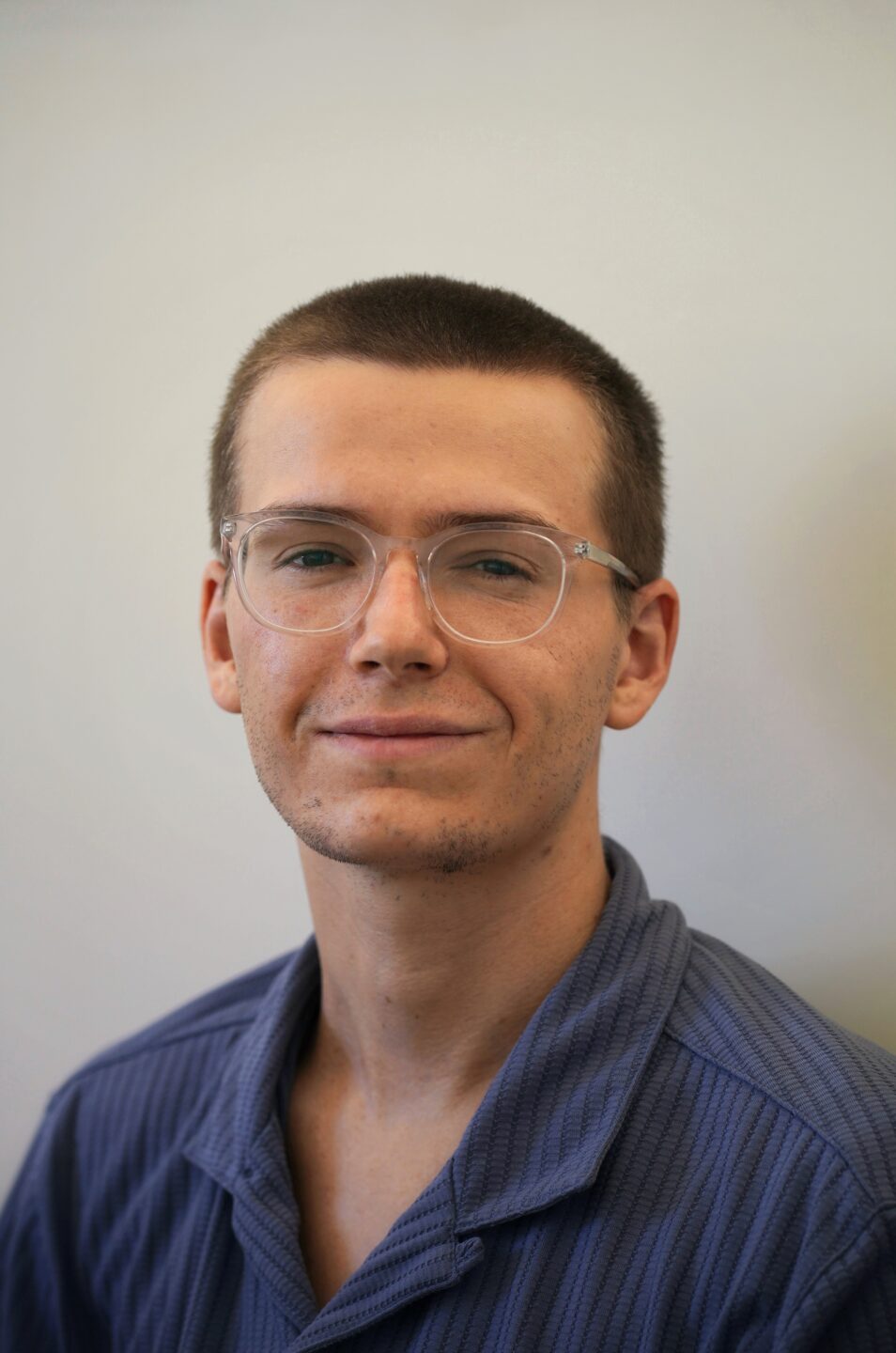 A photograph of Ned Lupson. He is smiling. His dark blonde hair is shaved. He is wearing glasses and a blue collared shirt.