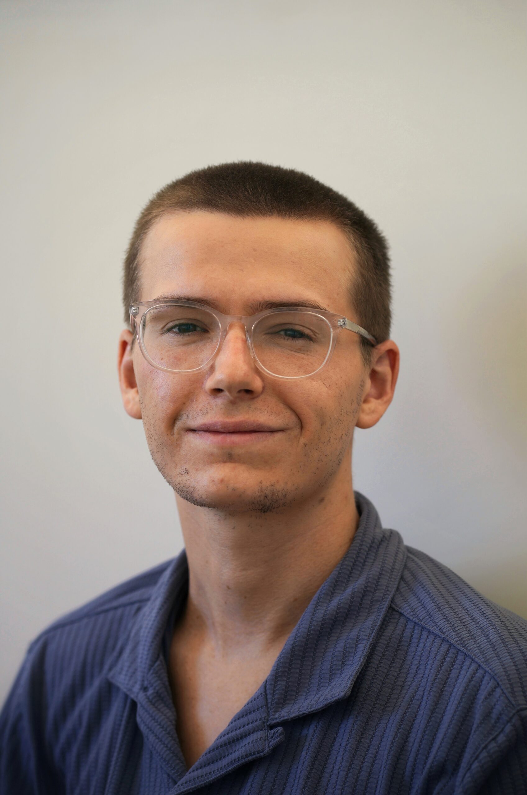 A photograph of Ned Lupson. He is smiling. His dark blonde hair is shaved. He is wearing glasses and a blue collared shirt.