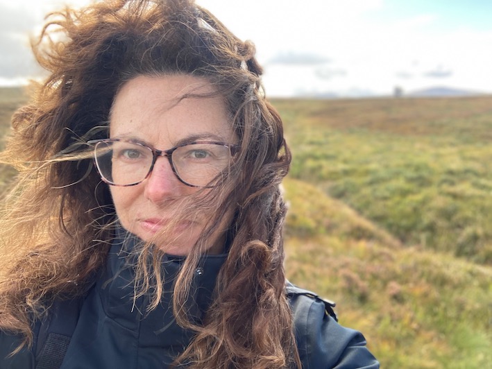 A portrait of Melissa Fagan in an outdoor setting. There are rolling hills behind her and her curly hair is blowing in the wind.