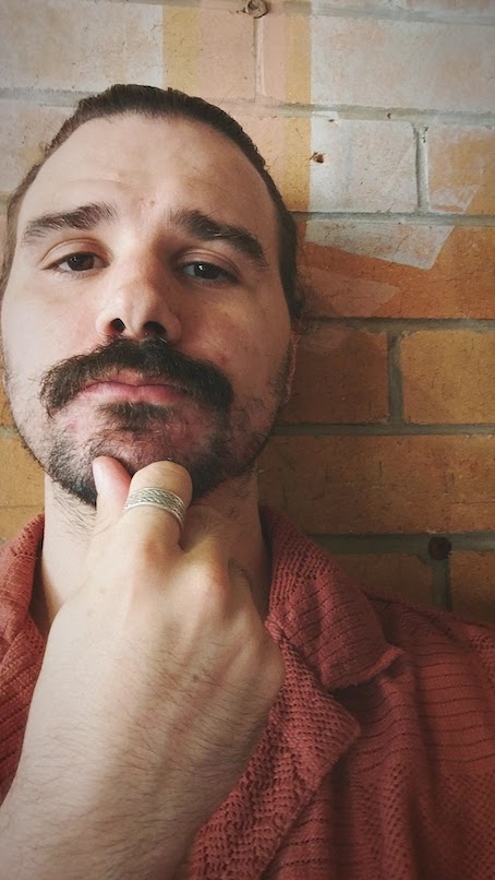 A portrait of the writer standing before a brick wall. He is wearing an orange shirt and hand his resting on his chin.