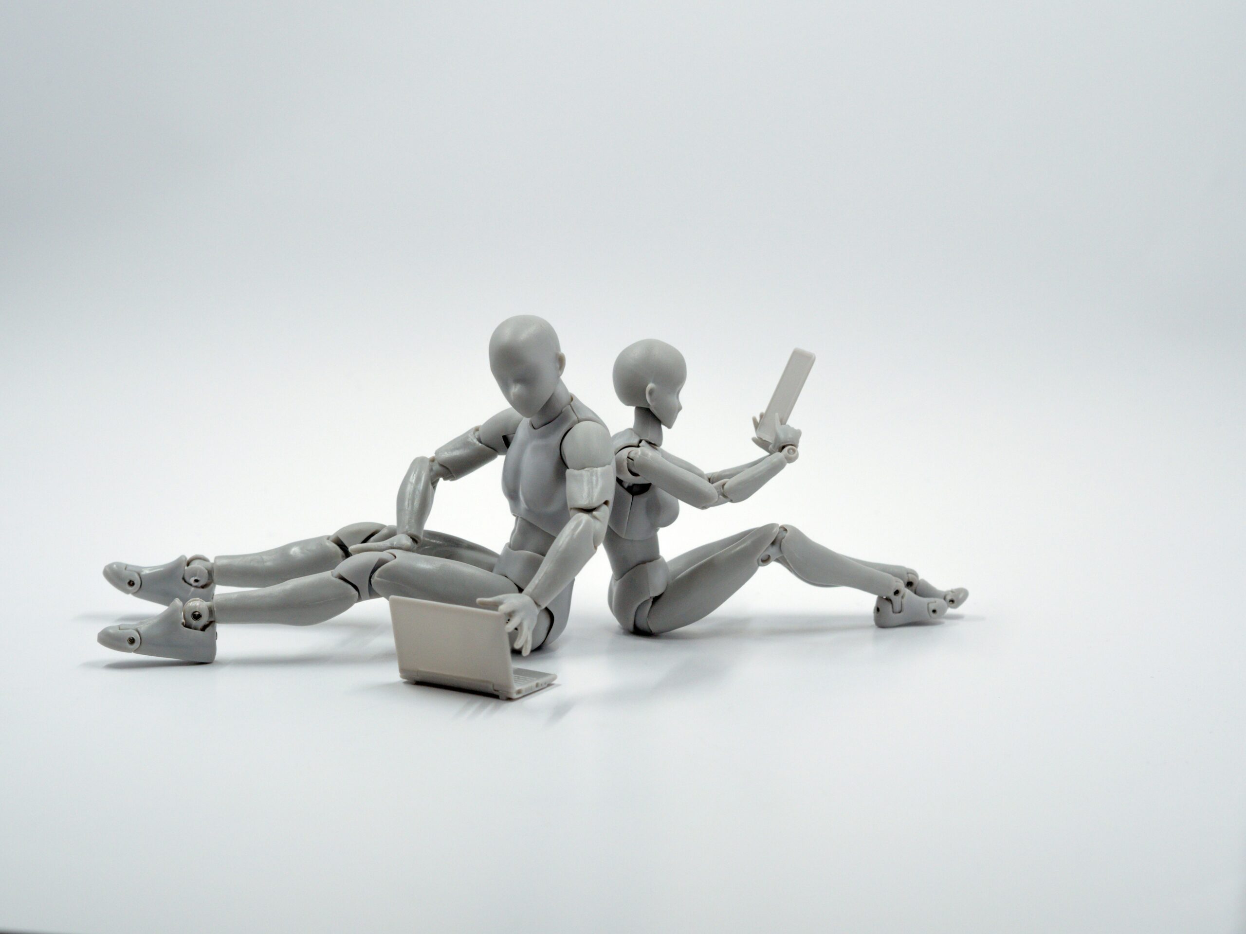 Two robots sit back to back. One is typing on a laptop and the other is scrolling on a smartphone.